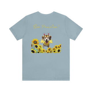 Bee Pawsitive Unisex Jersey Short Sleeve Tee shirt in Light Blue. Shown is back of shirt showcasing a dog dressed as as bee in a a field of sunflowers with the phrase "Bee Pawsitive!" above it. The front features the Bee Pawsitive Benefit Beagle Logo.