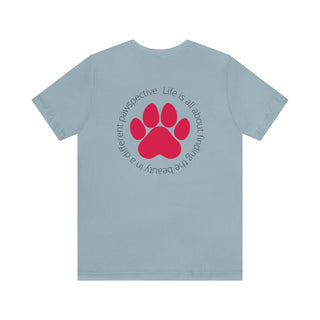 Different Pawspectives Unisex Jersey Short Sleeve Tee in Light Blue. Shown is the back of shirt featuring a large colorful pawprint with the the phrase "Life is all about finding the beauty in a different pawspective" circled around it. The Benefit Beagle Logo is located in the top corner on the front of shirt.