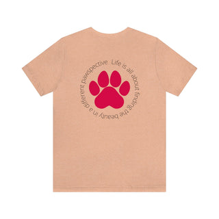 Different Pawspectives Unisex Jersey Short Sleeve Tee in Heather Peach. Shown is the back of shirt featuring a large colorful pawprint with the the phrase "Life is all about finding the beauty in a different pawspective" circled around it. The Benefit Beagle Logo is located in the top corner on the front of shirt.