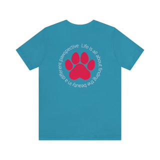 Different Pawspectives Unisex Jersey Short Sleeve Tee in Aqua. Shown is the back of shirt featuring a large colorful pawprint with the the phrase "Life is all about finding the beauty in a different pawspective" circled around it. The Benefit Beagle Logo is located in the top corner on the front of shirt.