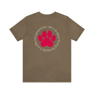 Different Pawspectives Unisex Jersey Short Sleeve Tee in Heather Olive. Shown is the back of shirt featuring a large colorful pawprint with the the phrase "Life is all about finding the beauty in a different pawspective" circled around it. The Benefit Beagle Logo is located in the top corner on the front of shirt.