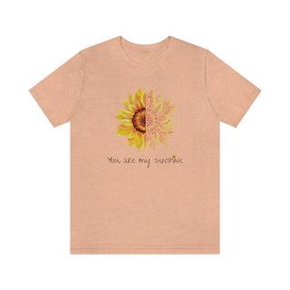 You are my Sunshine Unisex Jersey Short Sleeve Tee in Heather Peach. Shown is the front showcasing a sunflower which is split down the middle and half is made out of paw prints. Underneath is the phrase "You are my Sunshine" . Back of shirt features the Sunflower Benefit Beagle Logo.