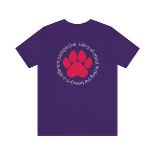 Different Pawspectives Unisex Jersey Short Sleeve Tee in Purple. Shown is the back of shirt featuring a large colorful pawprint with the the phrase "Life is all about finding the beauty in a different pawspective" circled around it. The Benefit Beagle Logo is located in the top corner on the front of shirt.