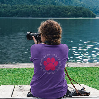 Different Pawspective Women's Softstyle Tee in Purple. Shown is the back of shirt featuring a large colorful pawprint with the the phrase "Life is all about finding the beauty in a different pawspective" circled around it. The Benefit Beagle Logo is located in the top corner on the front of shirt.