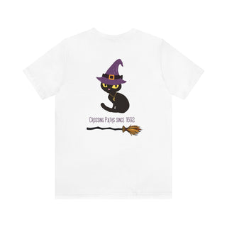 Witchy Cat Unisex Jersey Short Sleeve Tee. Back side shown in White with Wide Eyed Cartoon Cat wearing Purple Hat Standing over Broom. "Crossing Paths Since 1692". On the front of shirt is similar Witchy Benefit Beagle Logo. Purrfect for Halloween, or anytime!